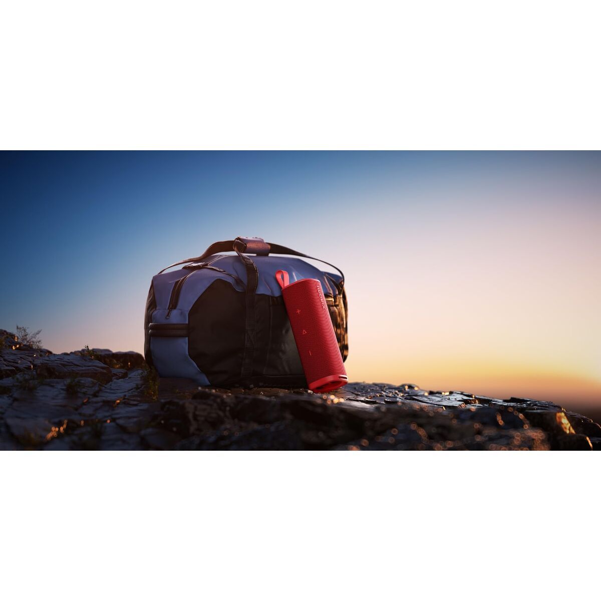 Rode Xiaomi Bluetooth luidspreker leunend tegen een zwarte tas bij zonsondergang.