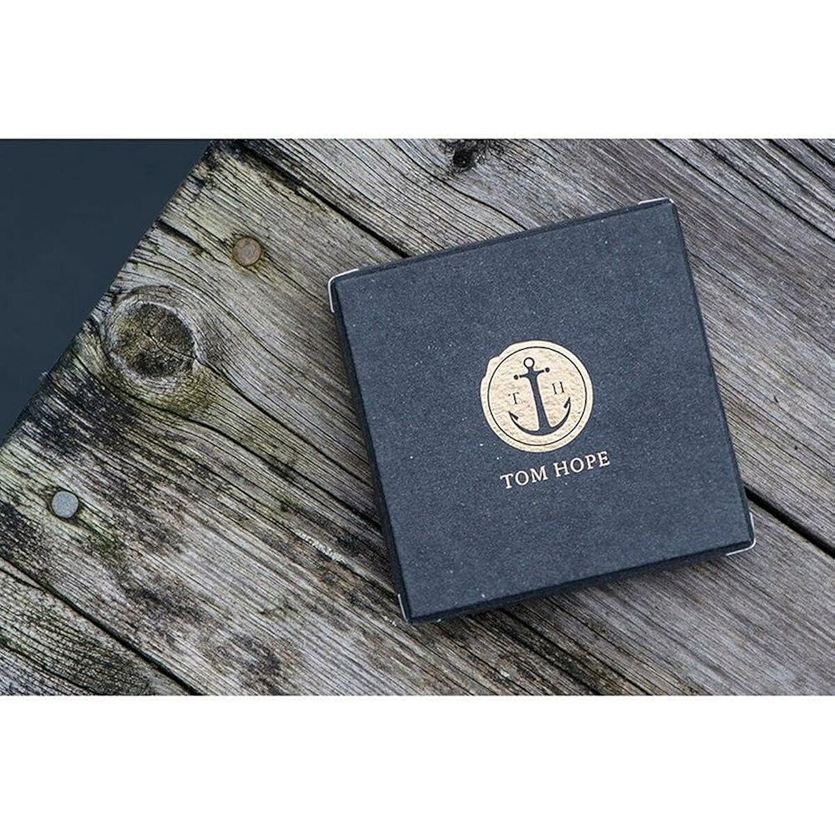 Zwarte merkdoos van Tom Hope met gouden logo op een houten ondergrond.